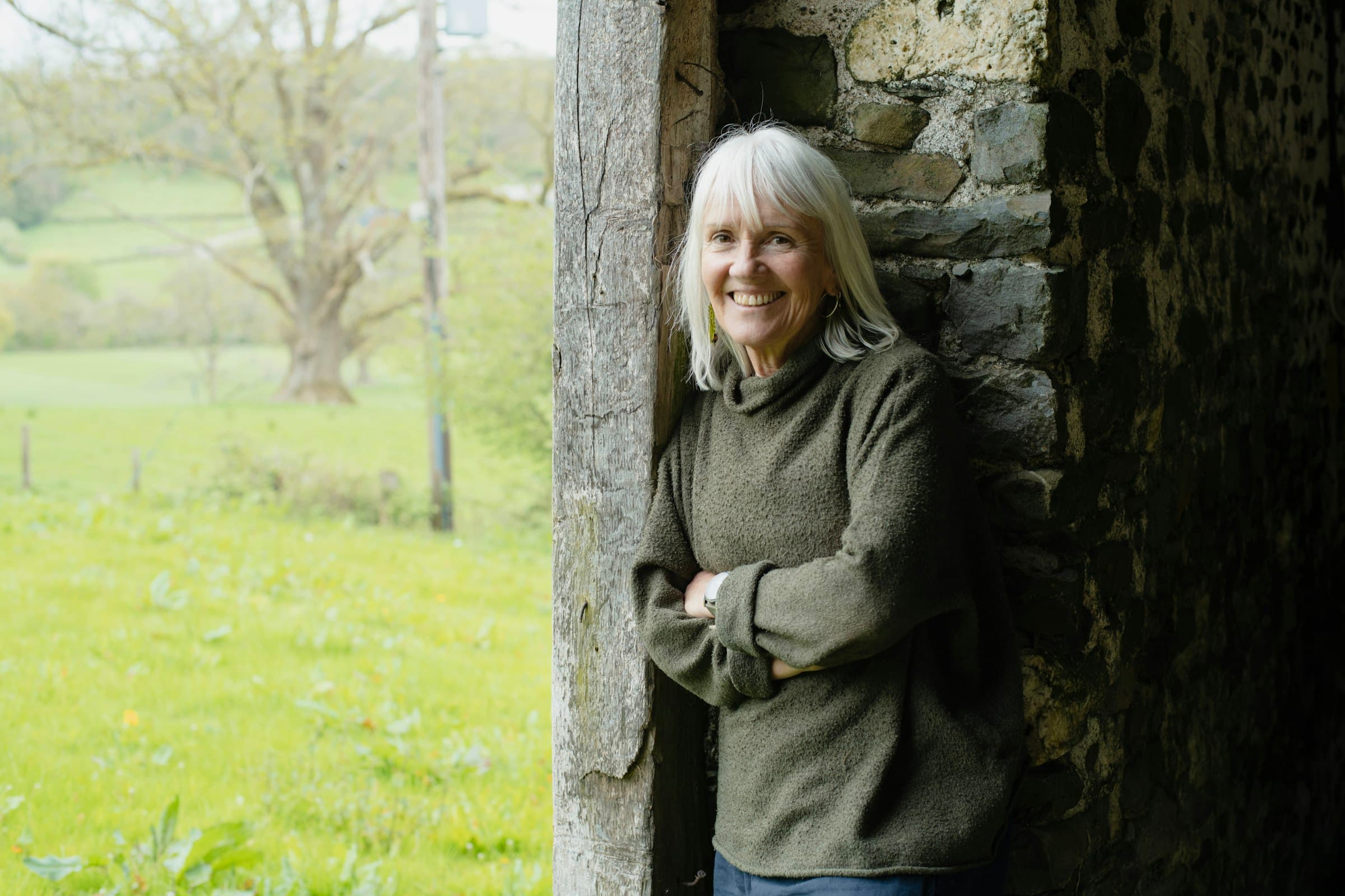 A woman with white hair in a green fleece leans against the stone doorway of a barn, arms folded, looking out over a green field in daylight.