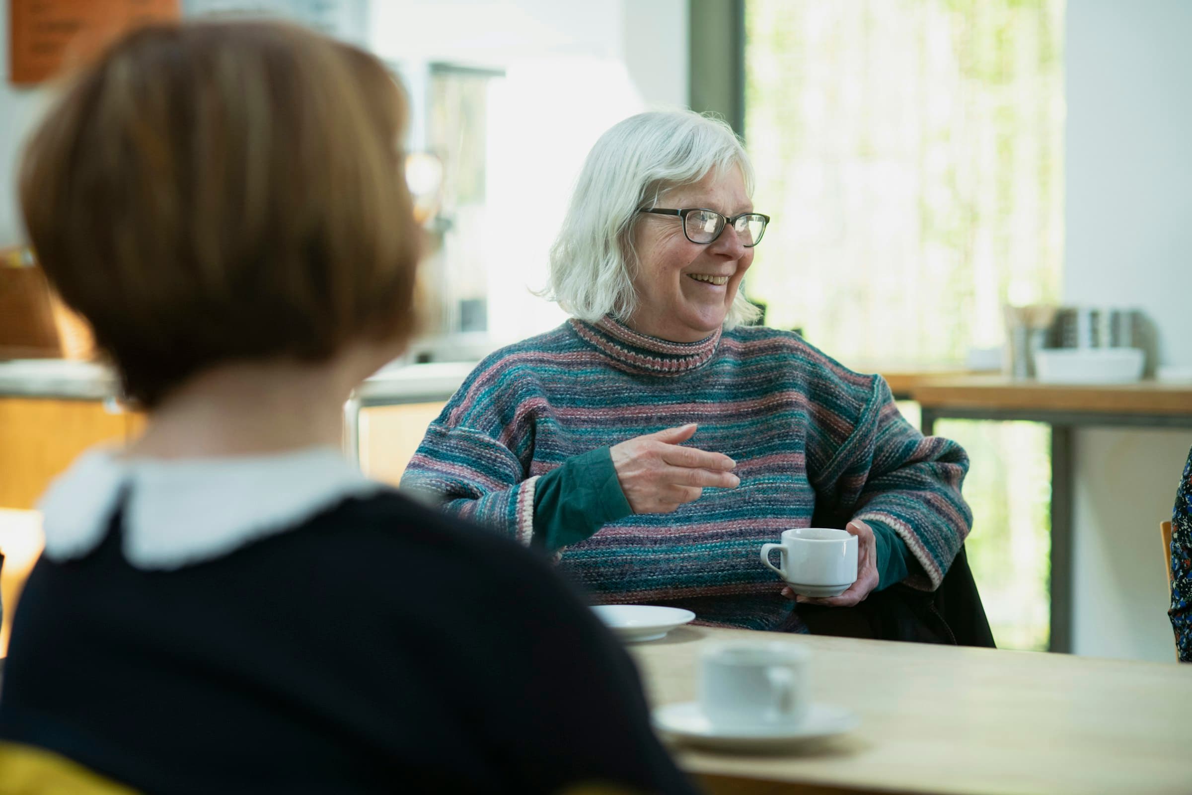 A white-haired woman in glasses and a striped jumper talks and gestures across a table over coffee, facing a younger woman seen from behind in the foreground.