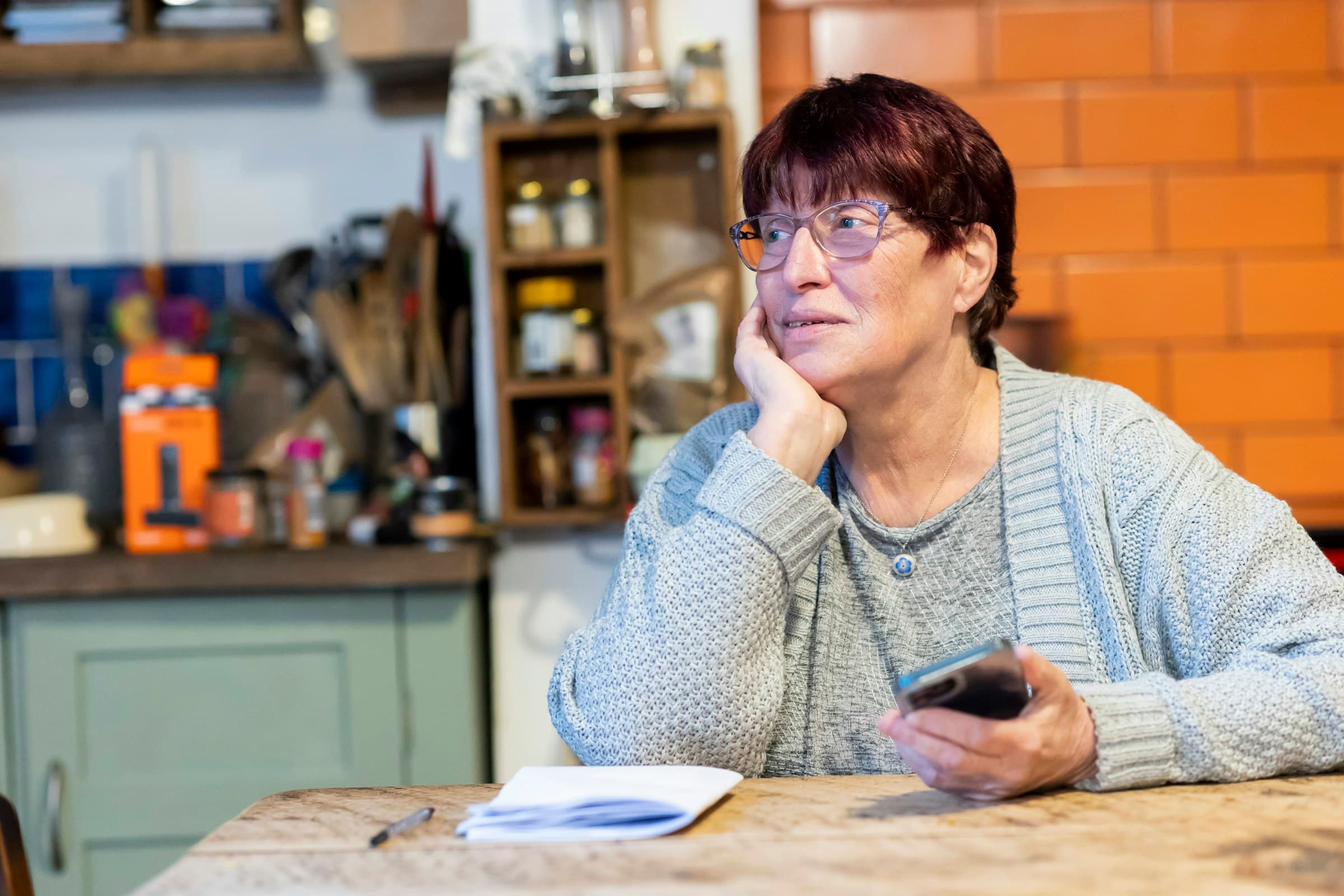 A woman in her sixties in a grey knitted cardigan sits at a wooden kitchen table in daylight, resting her chin on one hand and holding a mobile phone in the other, looking past the camera.