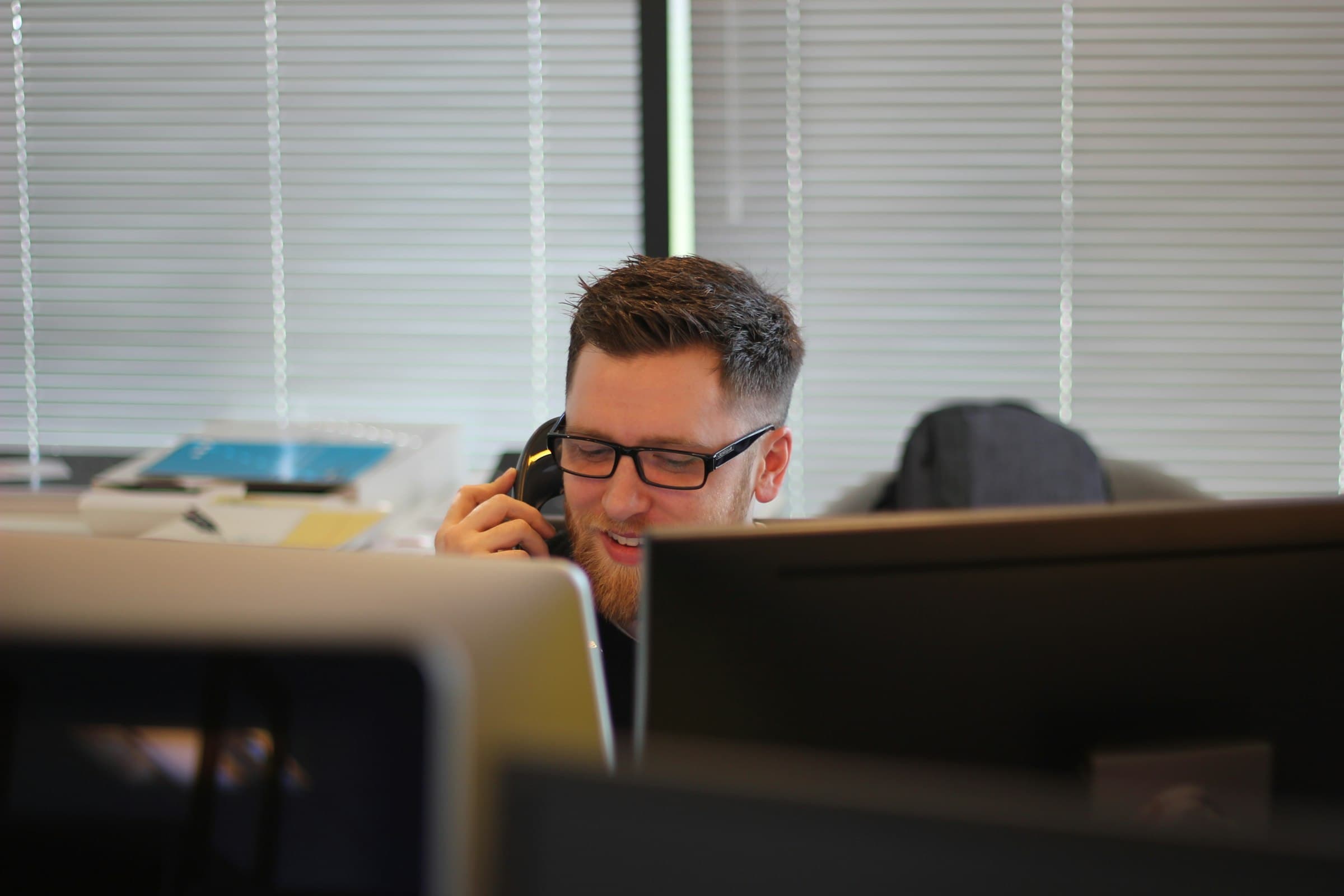 A man in glasses takes a call at his desk in a plain open-plan office, partly screened by monitors and window blinds.